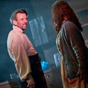 A man and woman stand facing each other in a dimly lit kitchen, with the man looking tense and the woman turned slightly away.