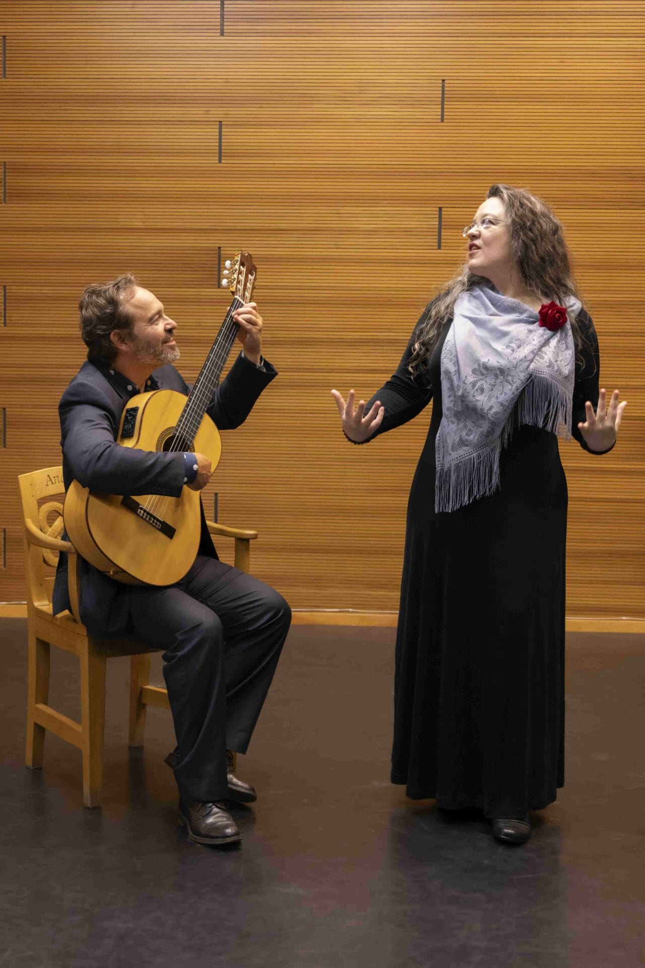 A man seated playing a classical guitar while a woman stands beside him singing, both performing in front of a wooden wall.
