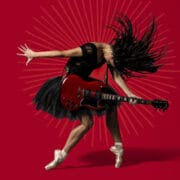 A woman wearing a black dress and pointe shoes plays an electric guitar while striking a dynamic pose against a red background with radiating lines.