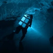 A scuba diver with dual tanks explores an underwater cave, illuminating the rocky surroundings with a flashlight—an adventure fit for the Ocean Film Festival.