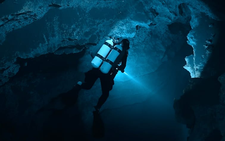 A scuba diver with dual tanks explores an underwater cave, illuminating the rocky surroundings with a flashlight—an adventure fit for the Ocean Film Festival.