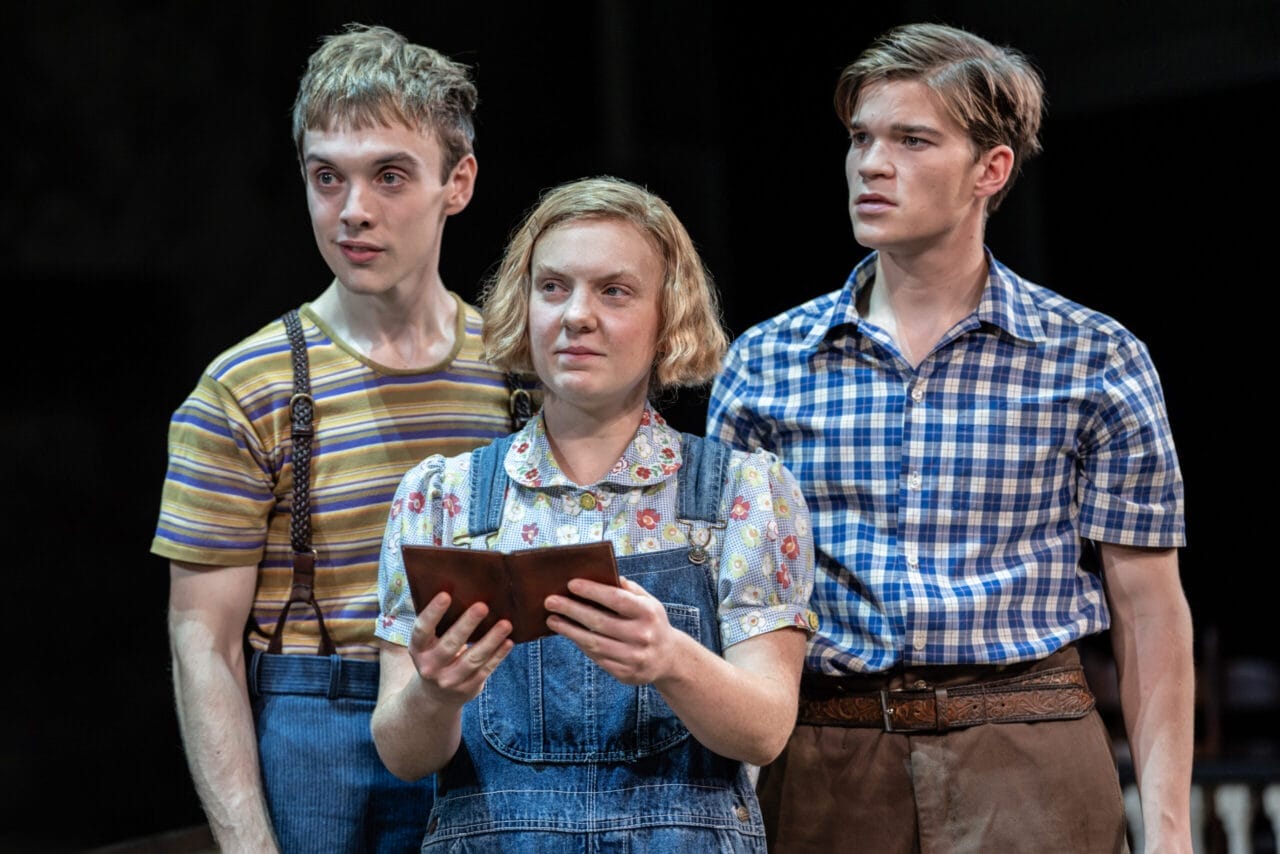 Three people stand close together; the woman in the center holds a brown book—perhaps a classic novel like To Kill a Mockingbird by Harper Lee—and wears overalls, while the two men beside her in striped and plaid shirts look forward.