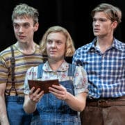 Three people stand close together; the woman in the center holds a brown book—perhaps a classic novel like To Kill a Mockingbird by Harper Lee—and wears overalls, while the two men beside her in striped and plaid shirts look forward.