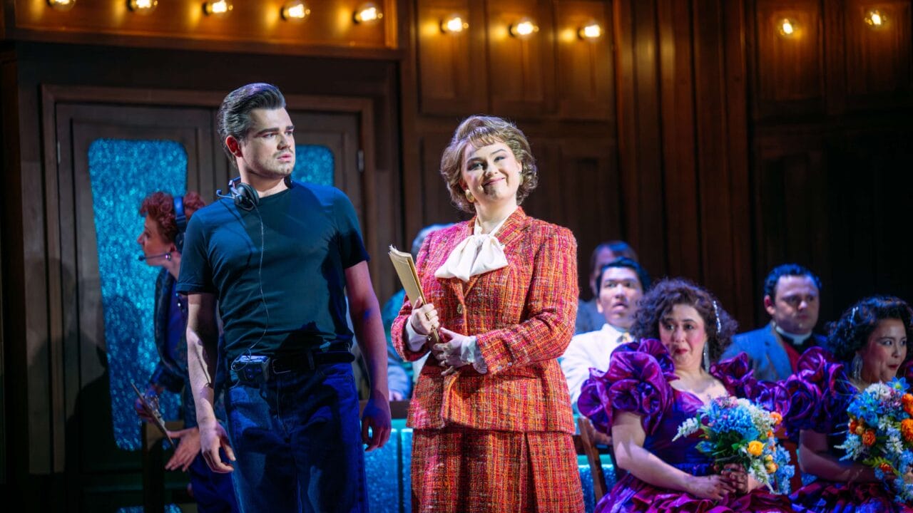 A woman in a red suit holds a folder and smiles on stage beside a man in black; others in colorful costumes sit behind them under warm stage lighting.