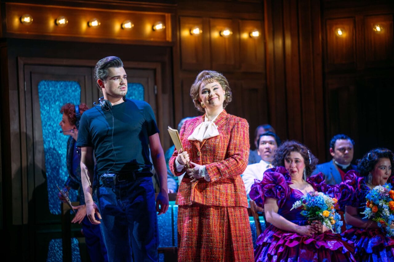 A woman in a red suit holds a folder and smiles on stage beside a man in black; others in colorful costumes sit behind them under warm stage lighting.
