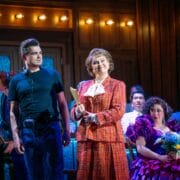 A woman in a red suit holds a folder and smiles on stage beside a man in black; others in colorful costumes sit behind them under warm stage lighting.