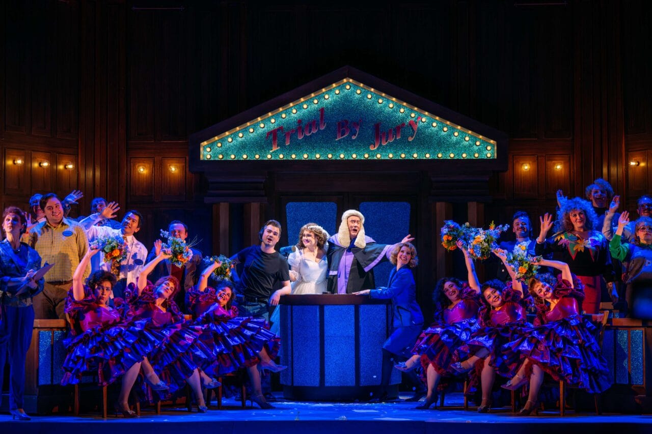 A theatrical performance depicts a lively courtroom scene with actors in colorful costumes, dancers in purple dresses, and a judge at the center under a sign reading "Trial By Jury.