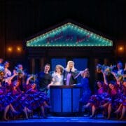 A theatrical performance depicts a lively courtroom scene with actors in colorful costumes, dancers in purple dresses, and a judge at the center under a sign reading "Trial By Jury.