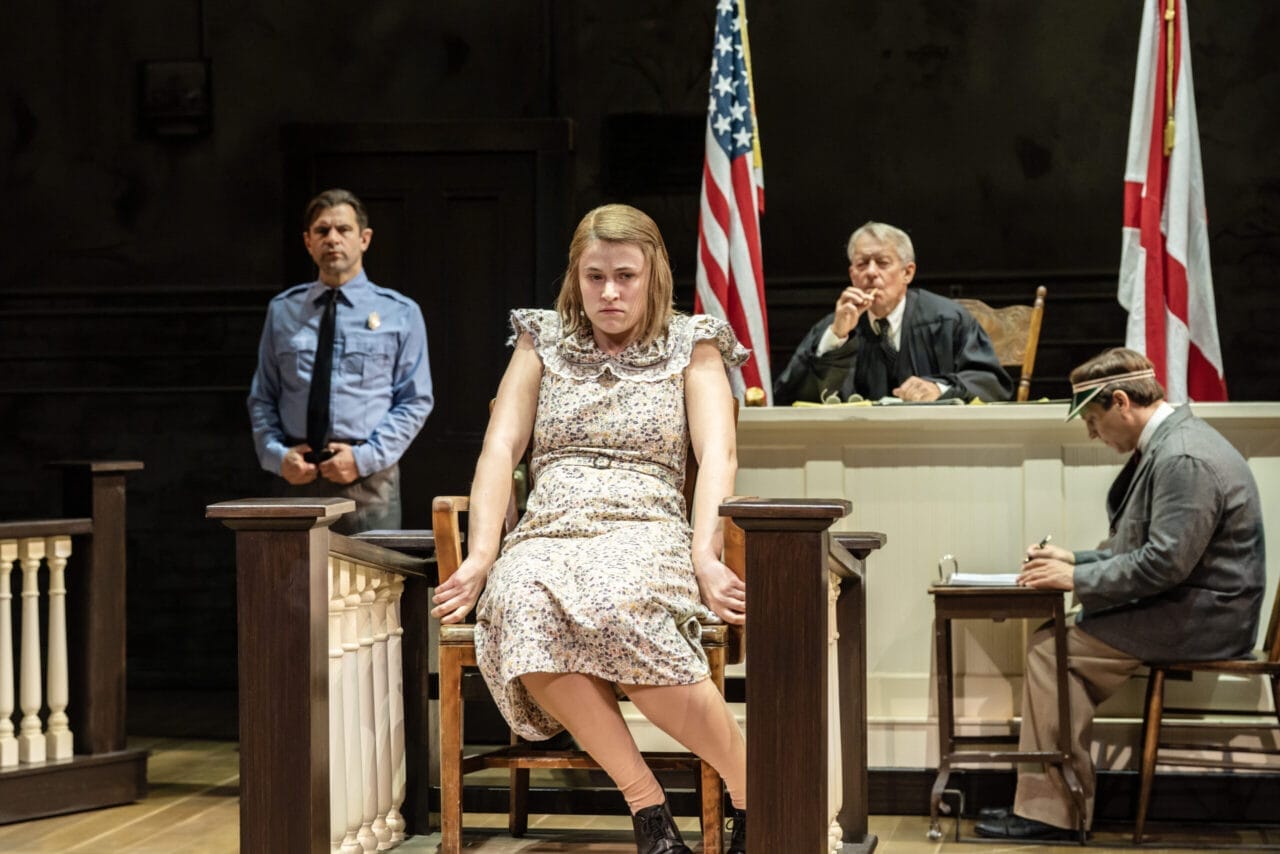 A woman in a floral dress sits on the witness stand in a courtroom, echoing a scene from Harper Lee's classic novel, To Kill a Mockingbird. The judge, police officer, two men, and US and Alabama flags are visible behind her.