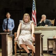 A woman in a floral dress sits on the witness stand in a courtroom, echoing a scene from Harper Lee's classic novel, To Kill a Mockingbird. The judge, police officer, two men, and US and Alabama flags are visible behind her.
