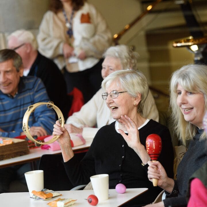 A group of older adults sits together at a table, some smiling and holding musical instruments like a tambourine and maracas, with refreshments and snacks nearby.