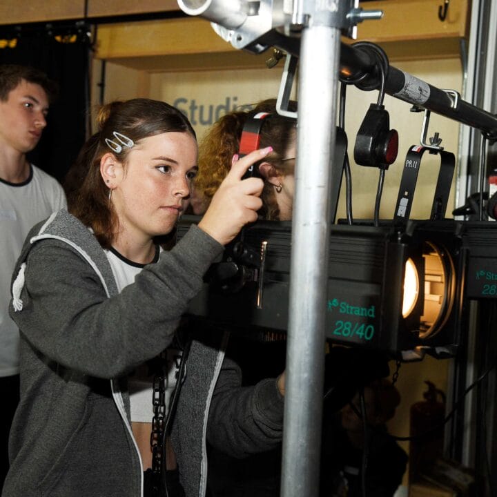 A girl adjusts stage lighting equipment while two other people observe in a backstage setting.