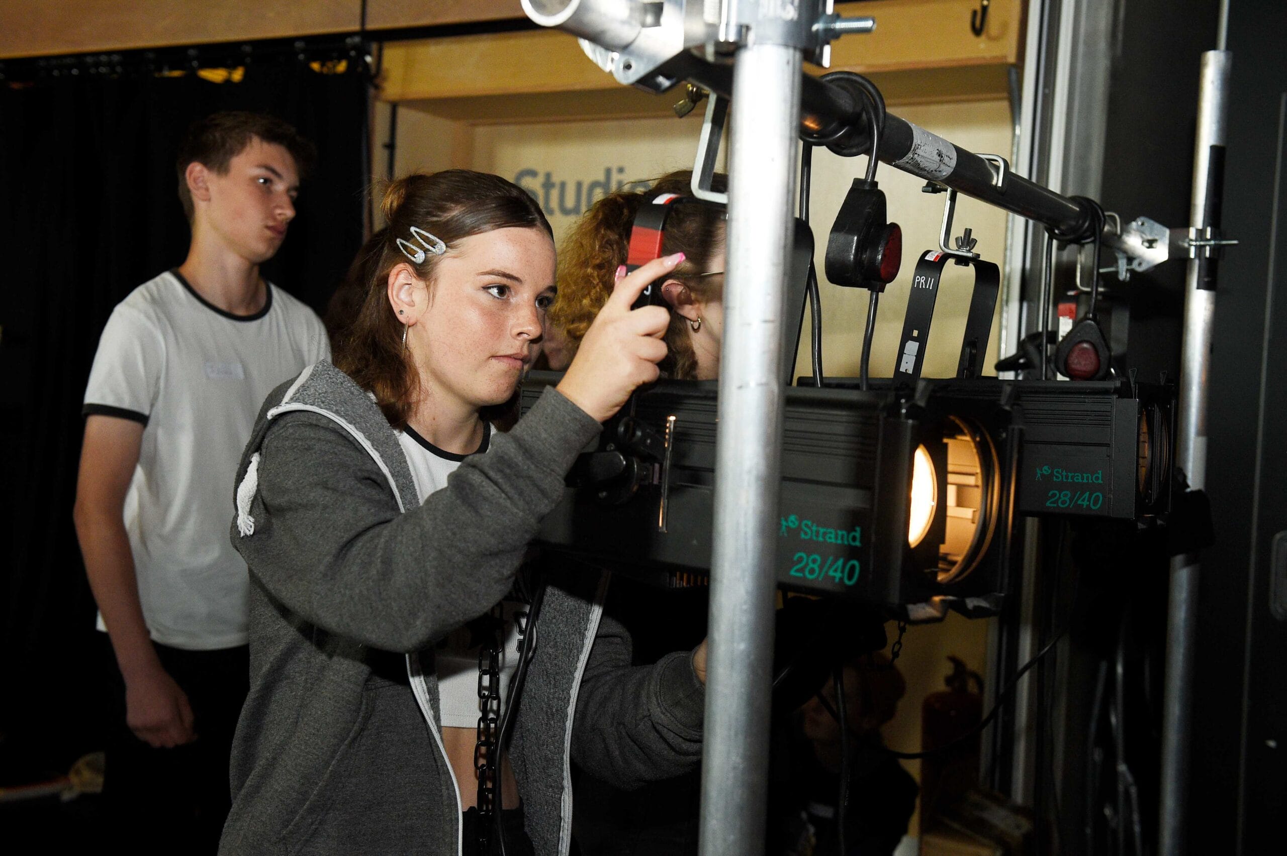 A girl adjusts stage lighting equipment while two other people observe in a backstage setting.
