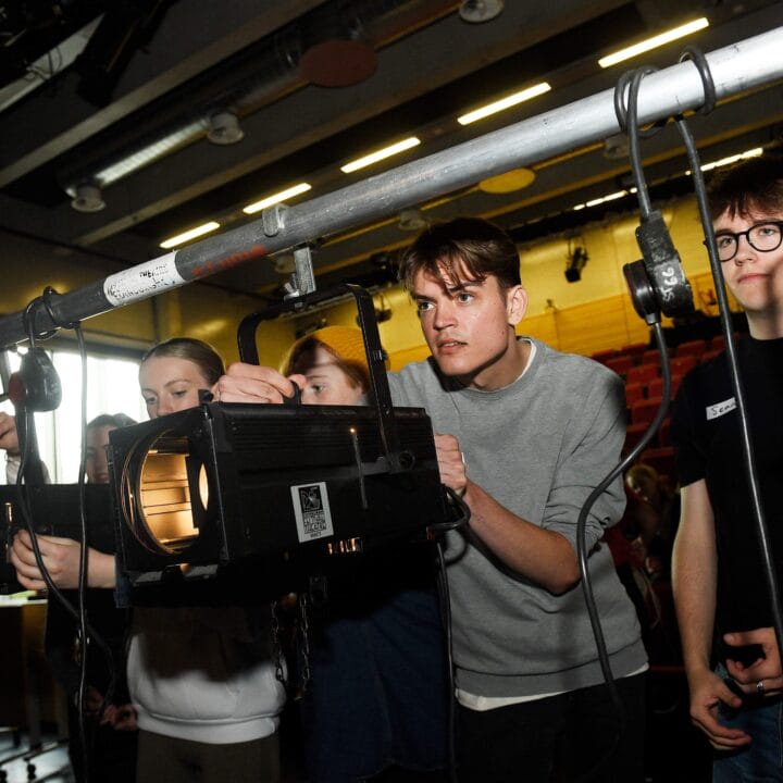 A group of people adjusting stage lighting equipment, focusing on positioning light fixtures on a metal rig in a theatre setting.