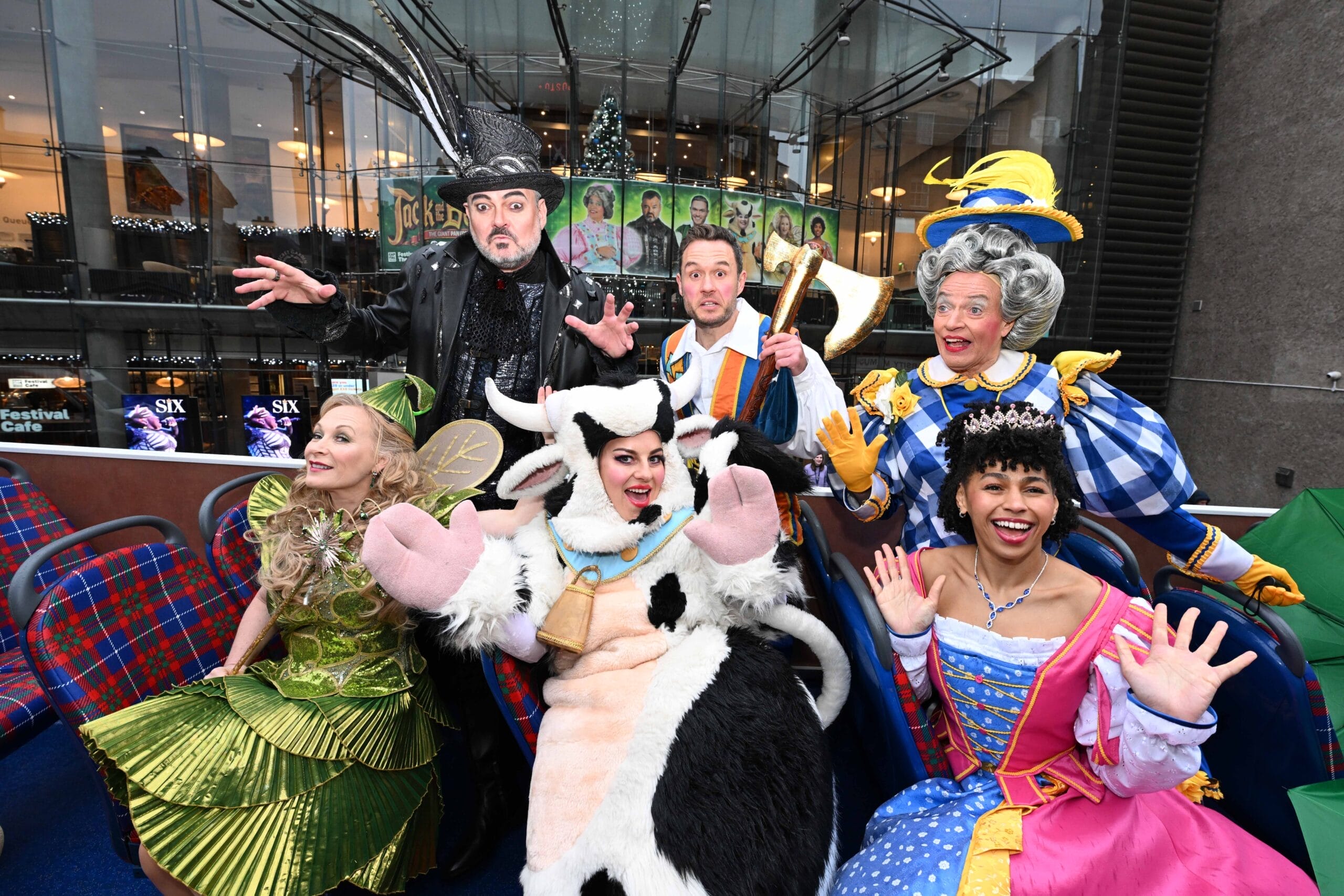 A group of six people in colorful, theatrical Jack and the Beanstalk costumes pose for a photo on a double-decker bus with a building and posters in the background.