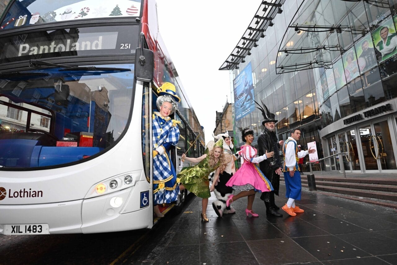 Six people in colorful pantomime costumes step off a city bus in an urban area, bringing a lively fairy tale vibe to the street, with a large glass building and bustling city life visible in the background.
