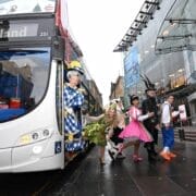 Six people in colorful pantomime costumes step off a city bus in an urban area, bringing a lively fairy tale vibe to the street, with a large glass building and bustling city life visible in the background.