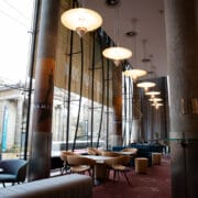 A bright and modern foyer area with round tables and chairs, large windows, hanging lights, and "Hamilton" banners, overlooking a classical building outside.