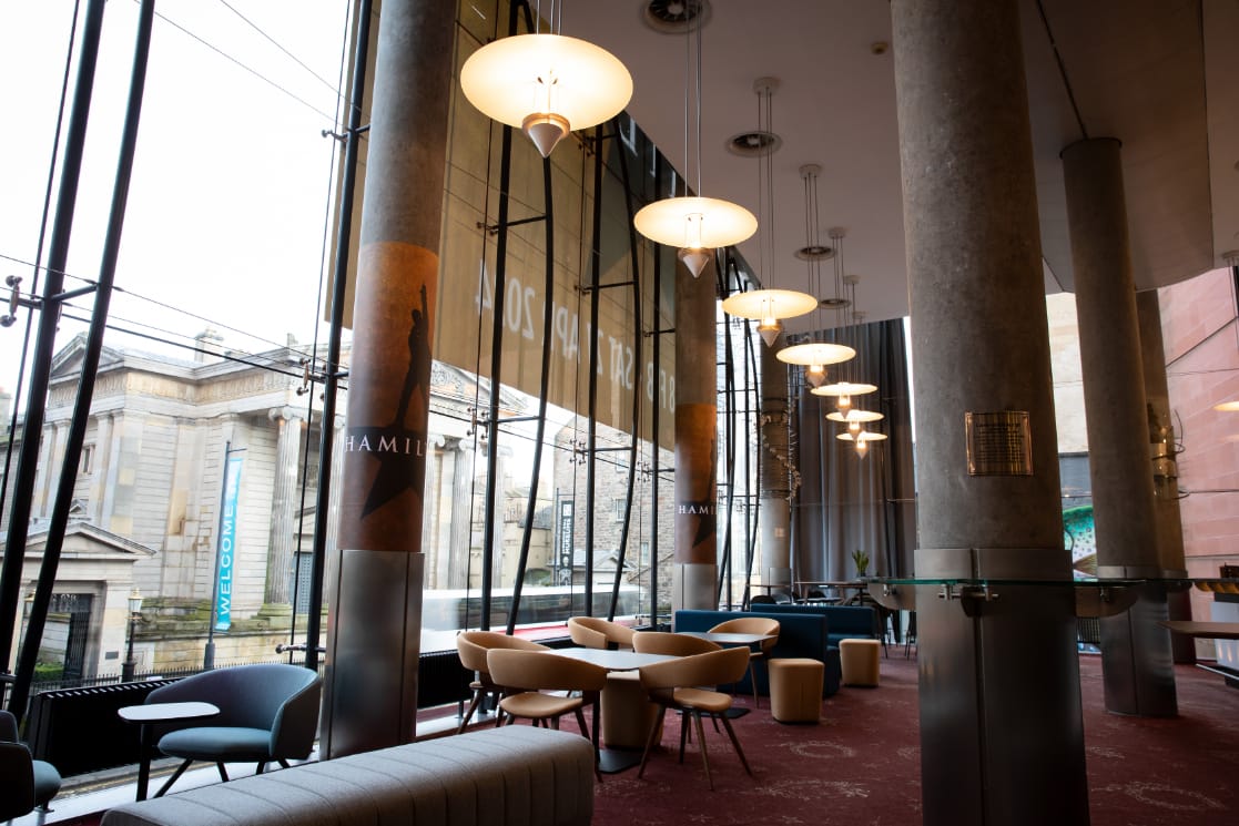A bright and modern foyer area with round tables and chairs, large windows, hanging lights, and "Hamilton" banners, overlooking a classical building outside.