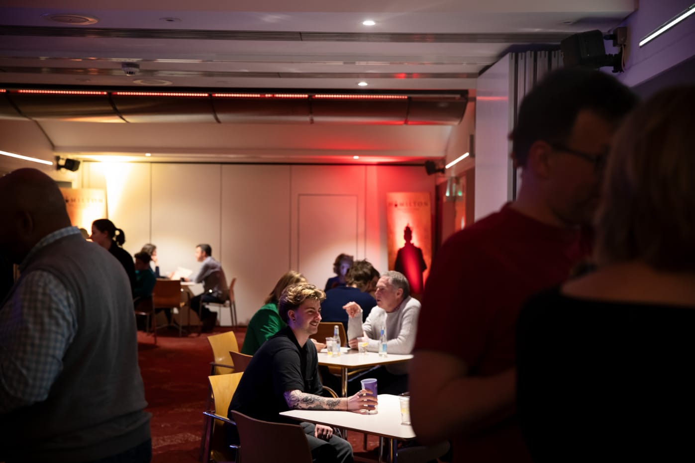 People sit and talk at tables in an atmospherically lit room with red carpeting and warm lighting on the walls. Some individuals stand and converse in the foreground.