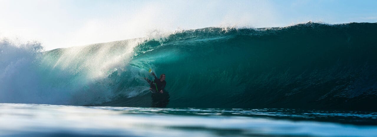 A surfer rides inside the barrel of a large, curling ocean wave under clear sky, illuminated by sunlight—capturing the spirit of adventure seen at the Ocean Film Festival.