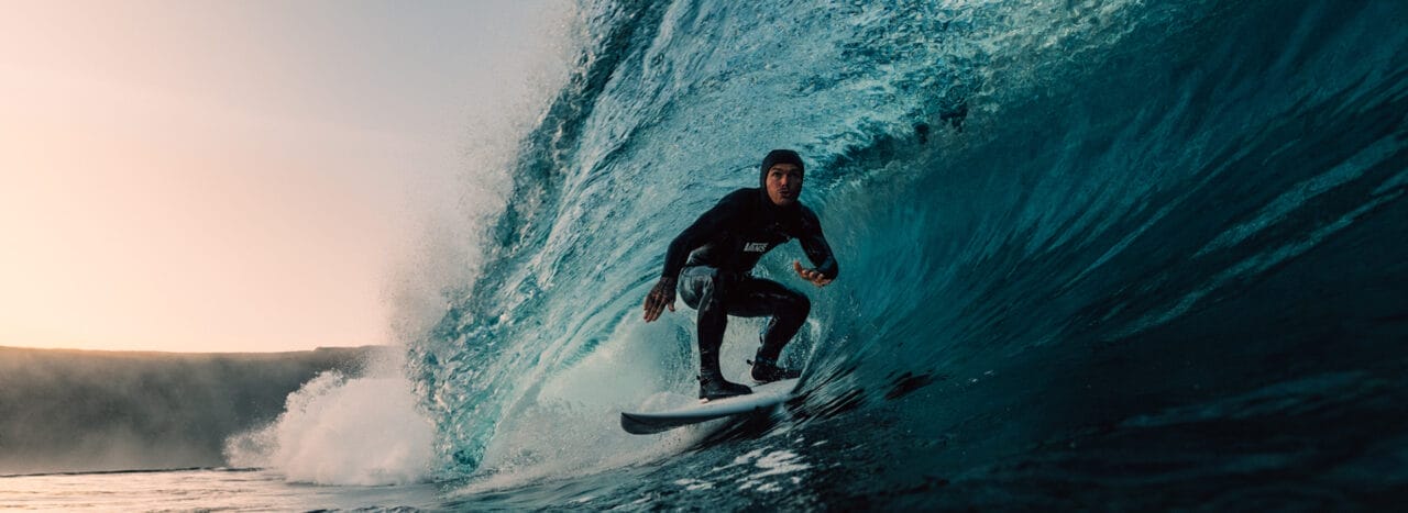 A surfer in a wetsuit rides inside a large, curling ocean wave near the shore at sunrise or sunset, capturing the spirit of adventure celebrated at the Ocean Film Festival.