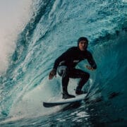 A surfer in a wetsuit rides inside a large, curling ocean wave near the shore at sunrise or sunset, capturing the spirit of adventure celebrated at the Ocean Film Festival.