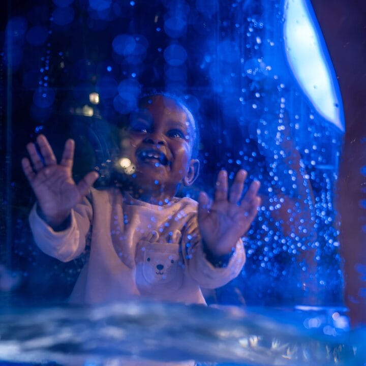 A smiling young child with hands pressed against a glass panel, illuminated by blue light with water droplets on the glass.
