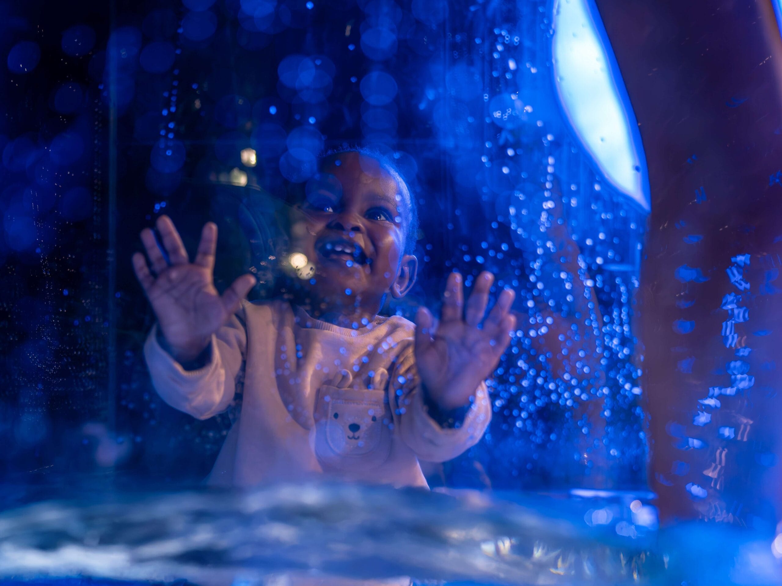 A smiling young child with hands pressed against a glass panel, illuminated by blue light with water droplets on the glass.