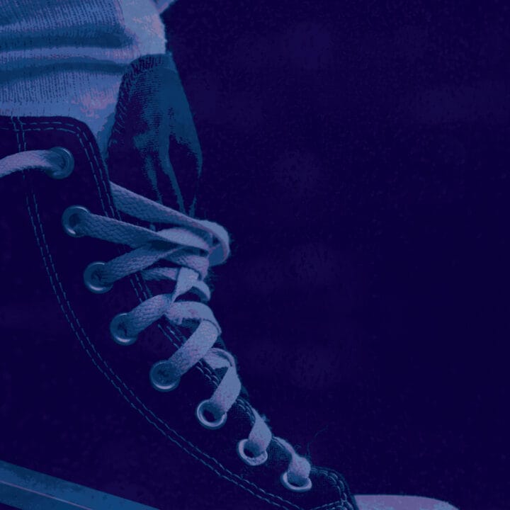 Close-up of a person wearing a lace-up sneaker with a blank circular tag attached to one eyelet, shown in blue and purple tones.