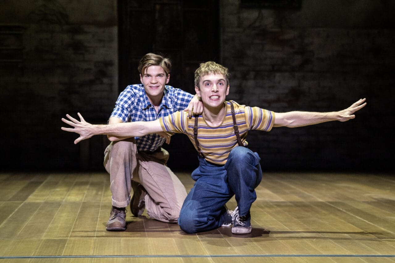 Two young men are on stage; one kneels and smiles, while the other crouches with arms outstretched like an airplane. Their casual, vintage-style clothing evokes the spirit of Harper Lee's classic novel, To Kill a Mockingbird.