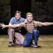 Two young men are on stage; one kneels and smiles, while the other crouches with arms outstretched like an airplane. Their casual, vintage-style clothing evokes the spirit of Harper Lee's classic novel, To Kill a Mockingbird.