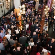 A large group of people are gathered indoors at a social event, standing and mingling near tables and a bar in the Festival Theatre, with tall columns and large windows.