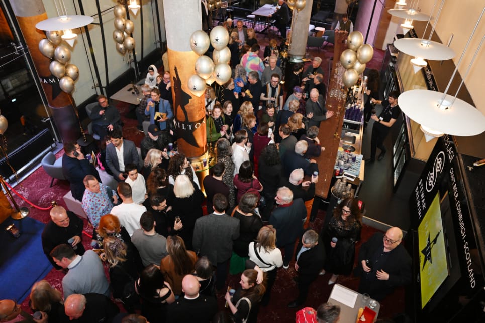 A large group of people are gathered indoors at a social event, standing and mingling near tables and a bar in the Festival Theatre, with tall columns and large windows.