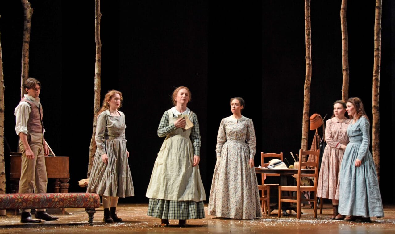 Six people in 19th-century attire stand on a stage set with chairs, a table, and tree trunks, suggesting a theatrical performance in a rustic setting.