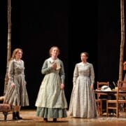 Six people in 19th-century attire stand on a stage set with chairs, a table, and tree trunks, suggesting a theatrical performance in a rustic setting.