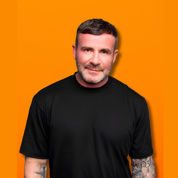 Gary Meikle, a man with short hair and tattoos on his arms, wearing a black shirt, stands against an orange background.