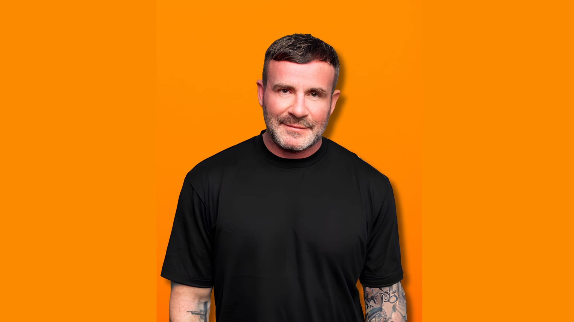 Gary Meikle, a man with short hair and tattoos on his arms, wearing a black shirt, stands against an orange background.
