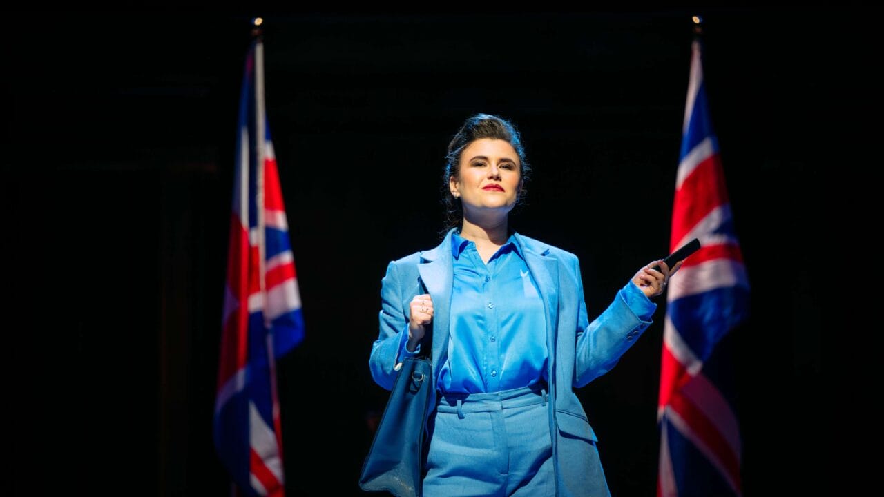 A person in a blue suit stands on stage between two British flags, holding a pen and looking ahead under bright stage lighting.