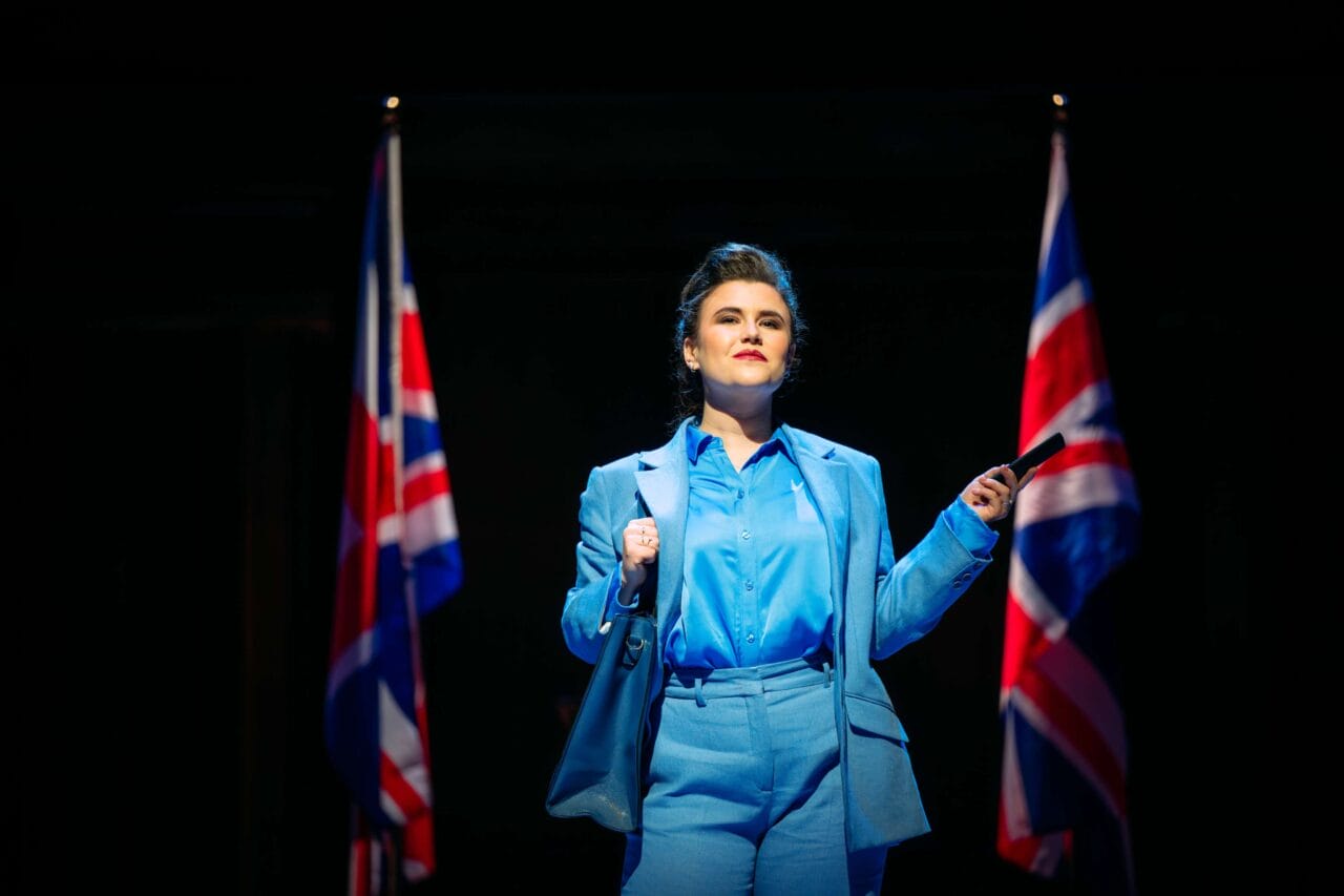 A person in a blue suit stands on stage between two British flags, holding a pen and looking ahead under bright stage lighting.