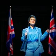 A person in a blue suit stands on stage between two British flags, holding a pen and looking ahead under bright stage lighting.