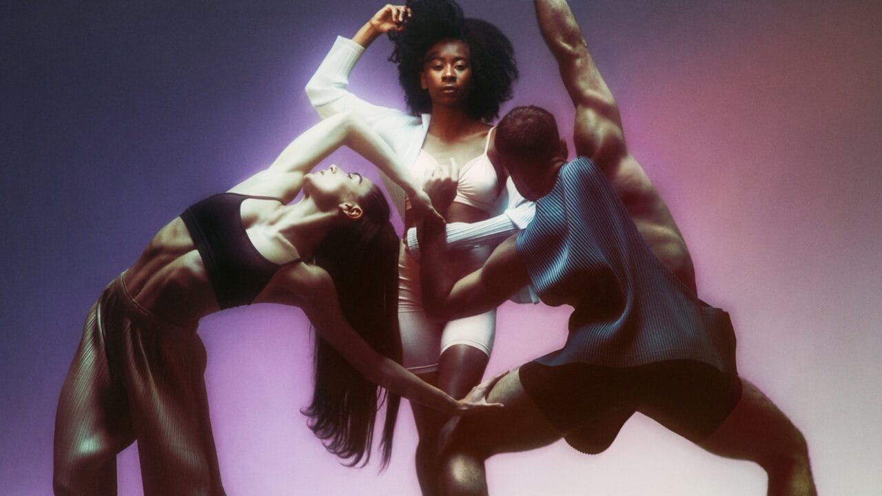 Three dancers pose in dynamic positions against a gradient pink and purple background, with one dancer standing upright and two bending and stretching in front.