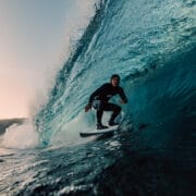 A surfer in a wetsuit rides inside a large blue wave near the shore at sunset, capturing the breathtaking spirit of the Ocean Film Festival.