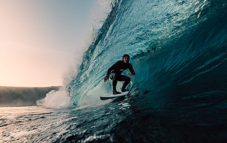 A surfer in a wetsuit rides inside a large blue wave near the shore at sunset, capturing the breathtaking spirit of the Ocean Film Festival.