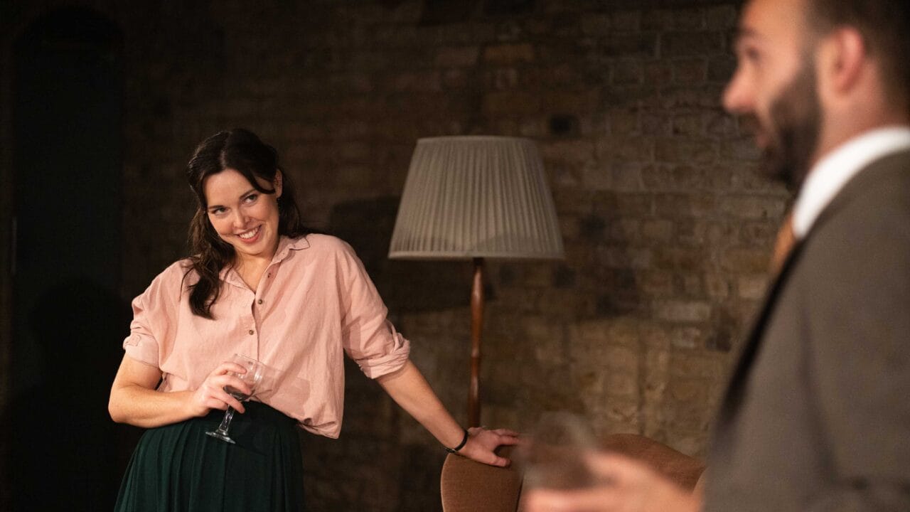 A woman holding a wine glass leans against a chair and smiles at a man in a suit who is also holding a glass; a floor lamp and brick wall are in the background.