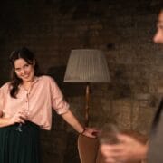 A woman holding a wine glass leans against a chair and smiles at a man in a suit who is also holding a glass; a floor lamp and brick wall are in the background.