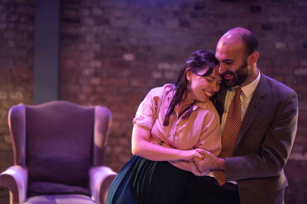 A smiling woman and man sit close together, holding hands and laughing, in front of a brick wall and an empty armchair.