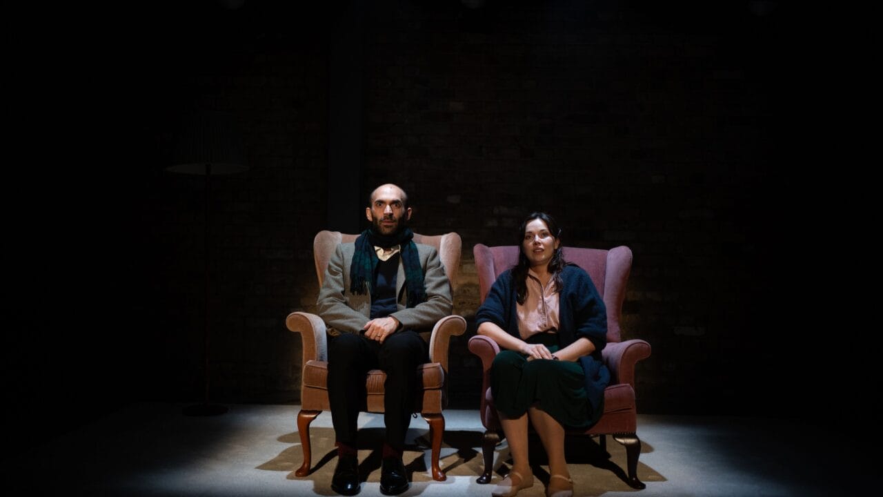 A man and a woman sit side by side in armchairs under a spotlight on a dark stage, facing forward with neutral expressions.