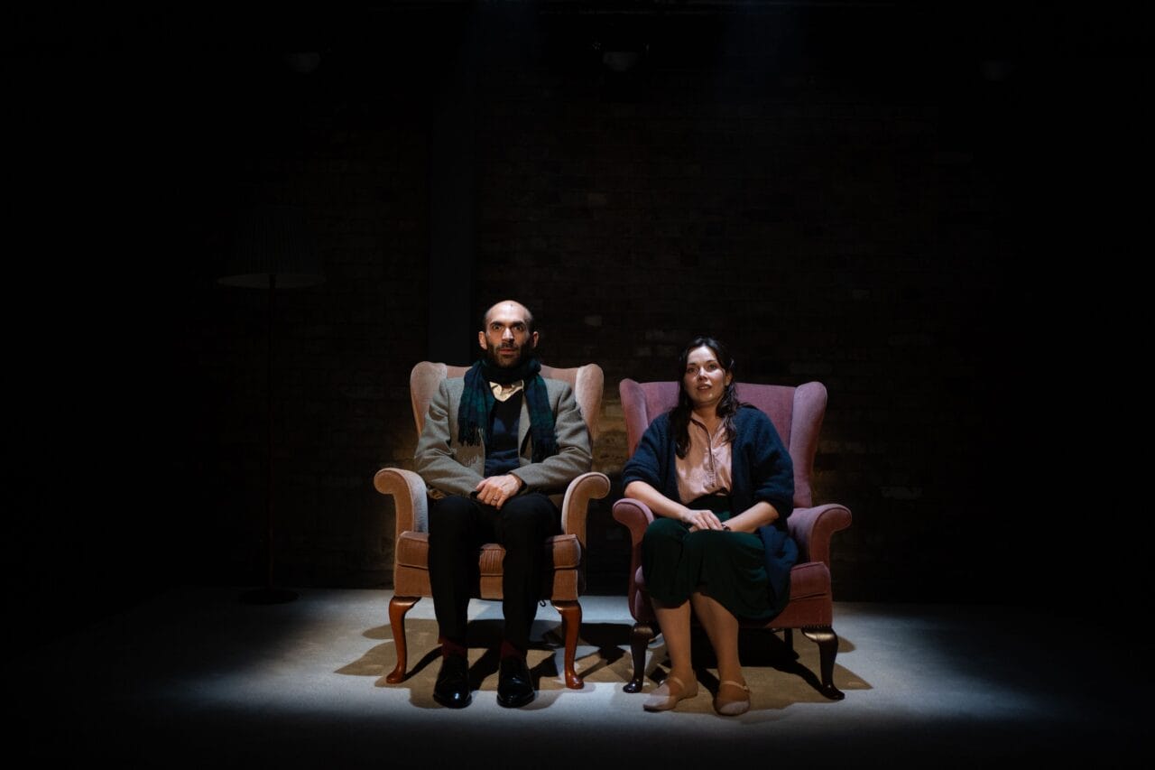 A man and a woman sit side by side in armchairs under a spotlight on a dark stage, facing forward with neutral expressions.