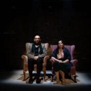 A man and a woman sit side by side in armchairs under a spotlight on a dark stage, facing forward with neutral expressions.